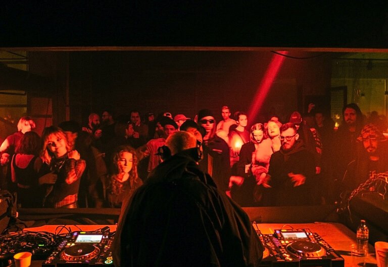 DJ performing at a dimly lit underground rave with a crowd of people dancing.