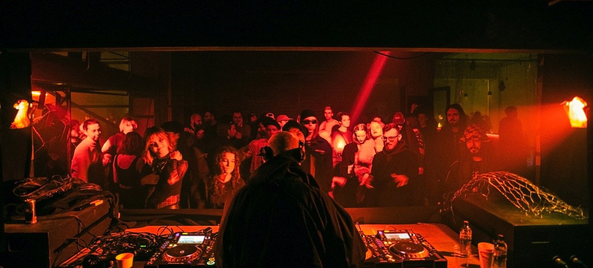 DJ performing at a dimly lit underground rave with a crowd of people dancing.